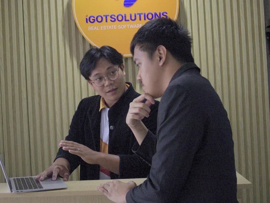 Two iGotSolutions consultants reviewing a real estate software workflow on a laptop during a client session at the company's Cebu office, beneath the iGotSolutions Real Estate Software sign